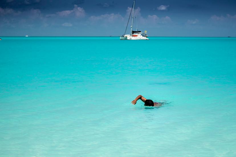 Man swimming to shore in perfectly clear turquoise-colored water. A catamaran sailboat at anchor is in the background.