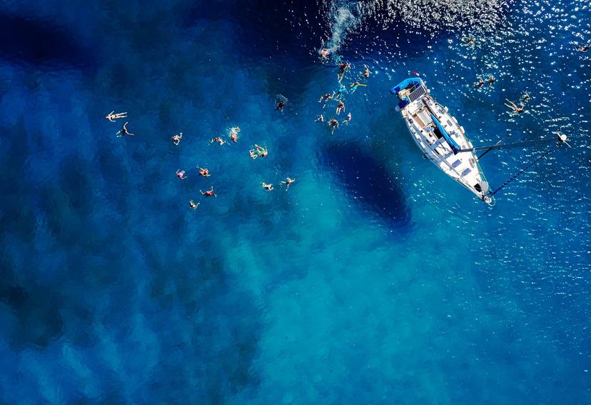 Aerial of a group of people swimming in blue water near a sailing boat in Barbados.