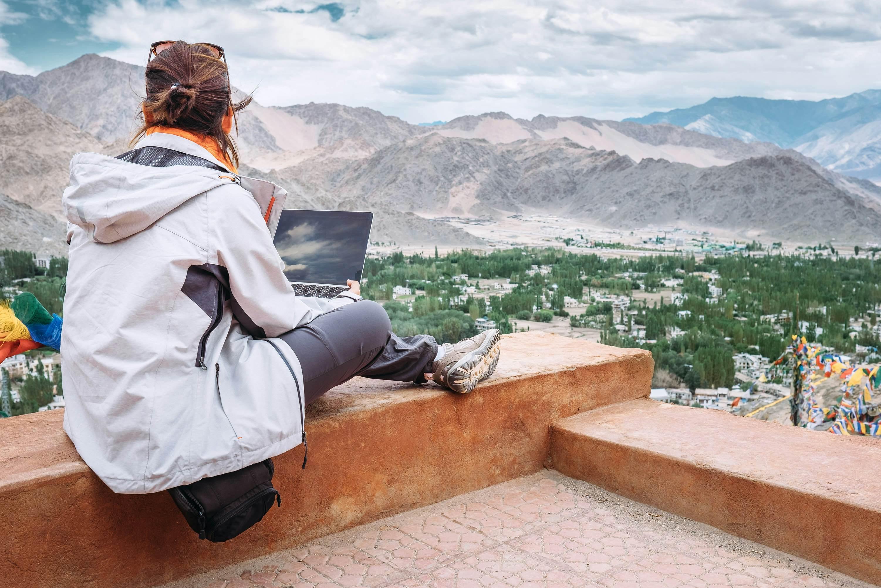 Female traveler with a laptop sitting at a viewpoint above a mountainous valley in India.  