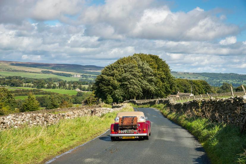 Little red car driving near Aysgarth in the Yorkshire Dales on a sunny September day. Bolton castle seen in the far distance.