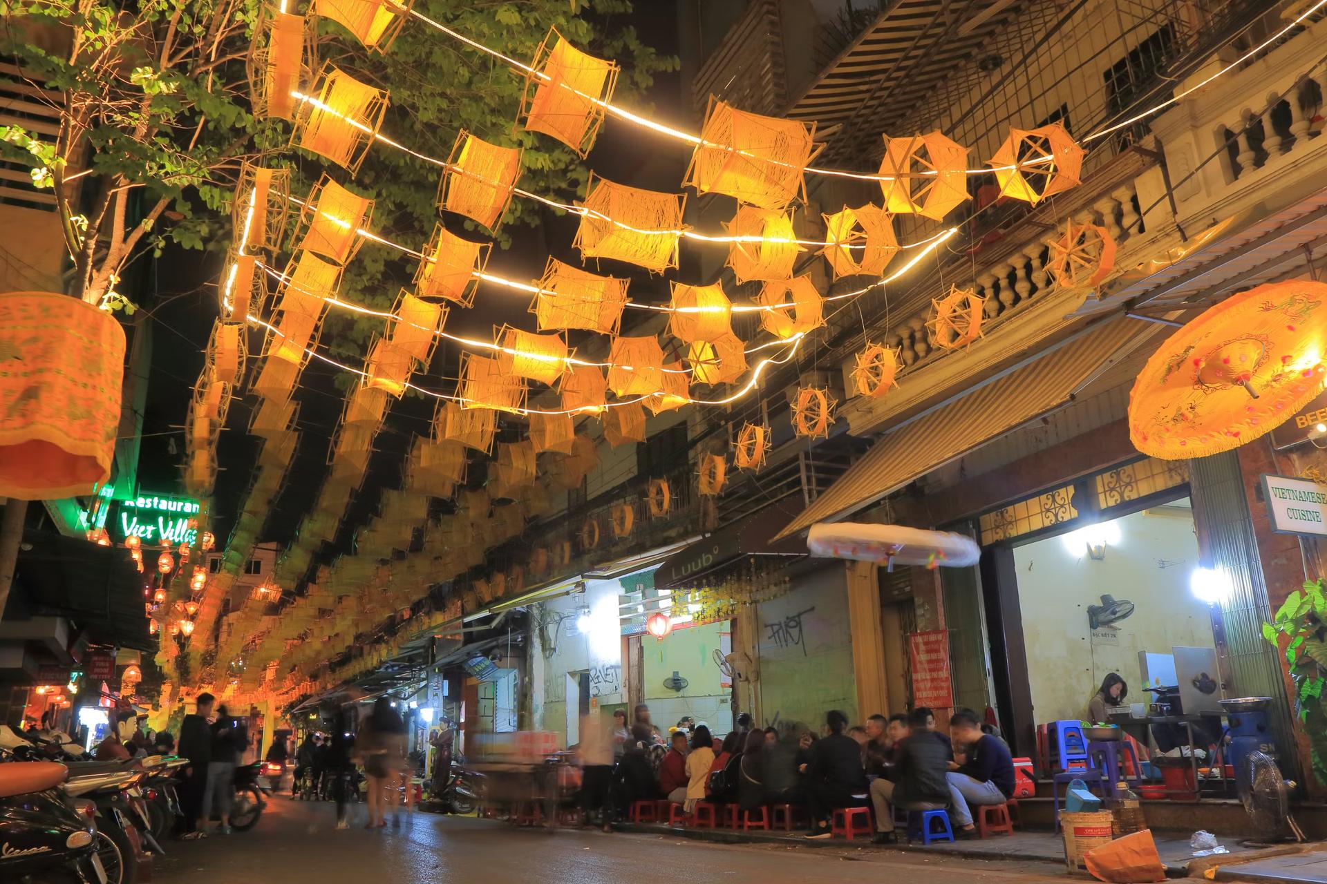 Folk sitter utanför barer och restauranger i Hanois gamla kvarter.  Det är natt och bord och stolar har ställts ut på vägen utanför verksamheterna.