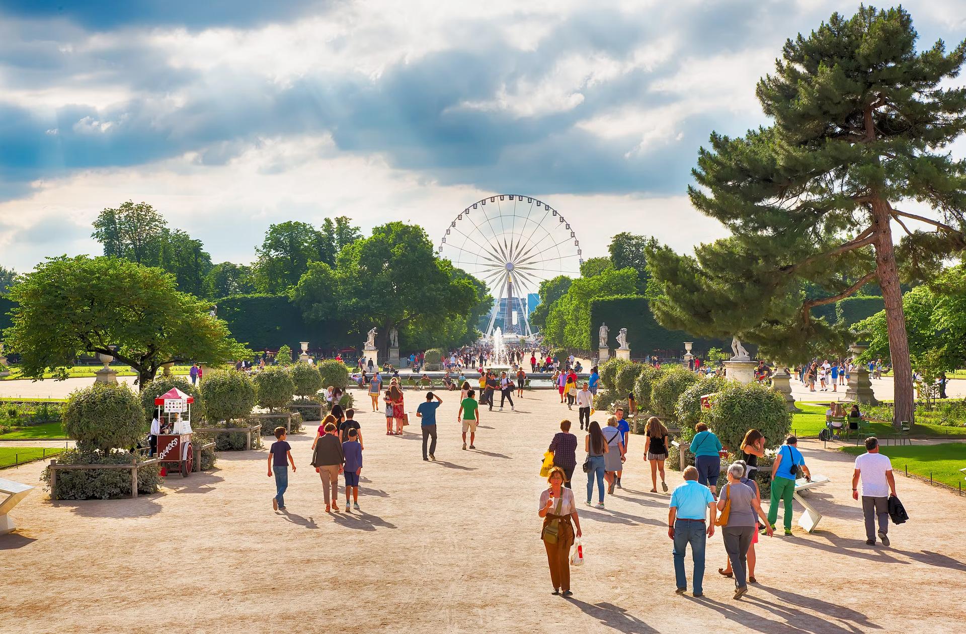 Separate groups of people walk down a wide pathway through a garden towards a large Ferris wheel in the distance 