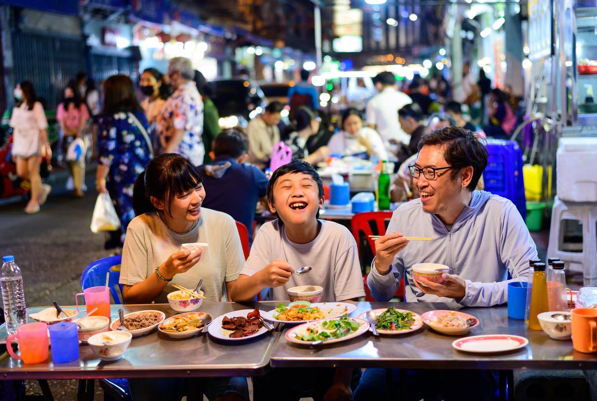 Den asiatiska familjen njuter av att äta mat på en gatumatrestaurang med folkmassa på Yaowarat Road, Bangkok