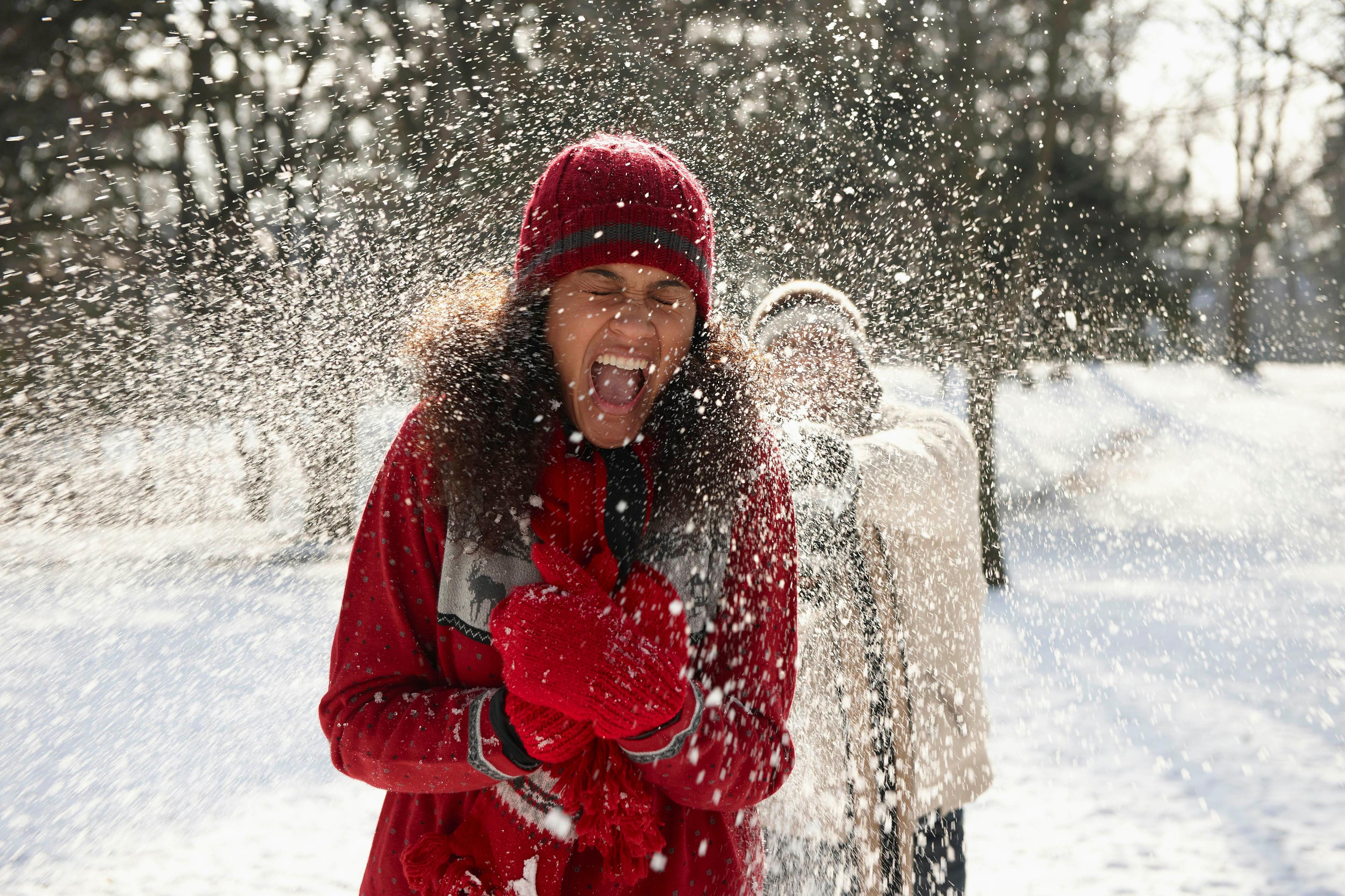A woman wearing a bright red coat and hat, screams as she's hit with a snowball from behind. 