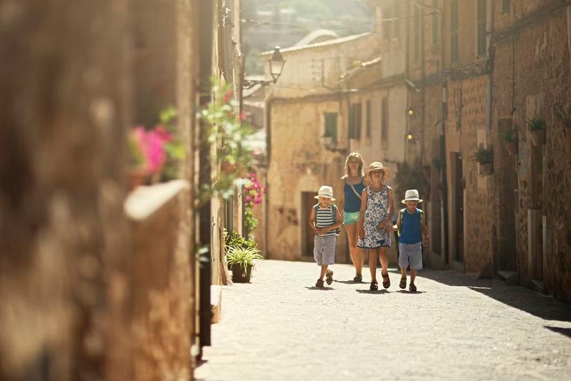 Mother and three kids walking in the beautiful mediterranean town. Little boys are wearing backpacks and hats. Theirs sister is wearing sundress. Sunny summer day. Mallorca, Spain, Valdemossa.