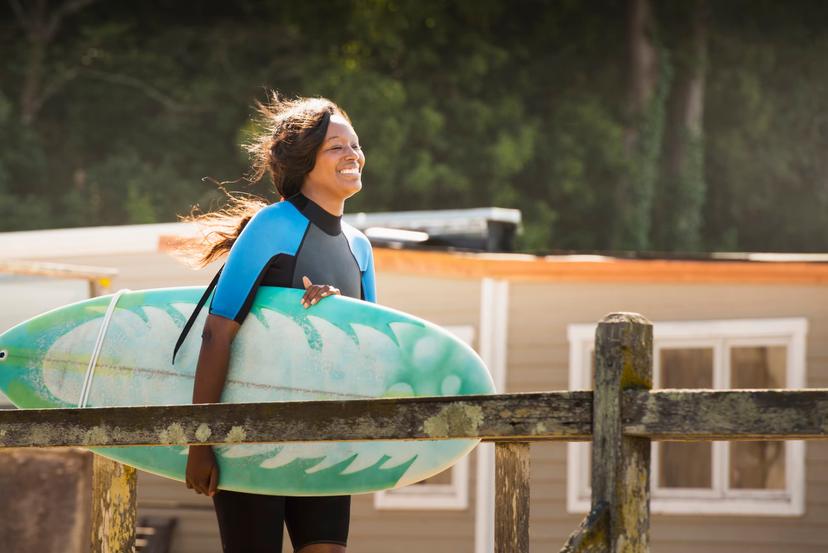 Smiling African-American woman walks down a pier in a wetsuit carrying a surfboard
