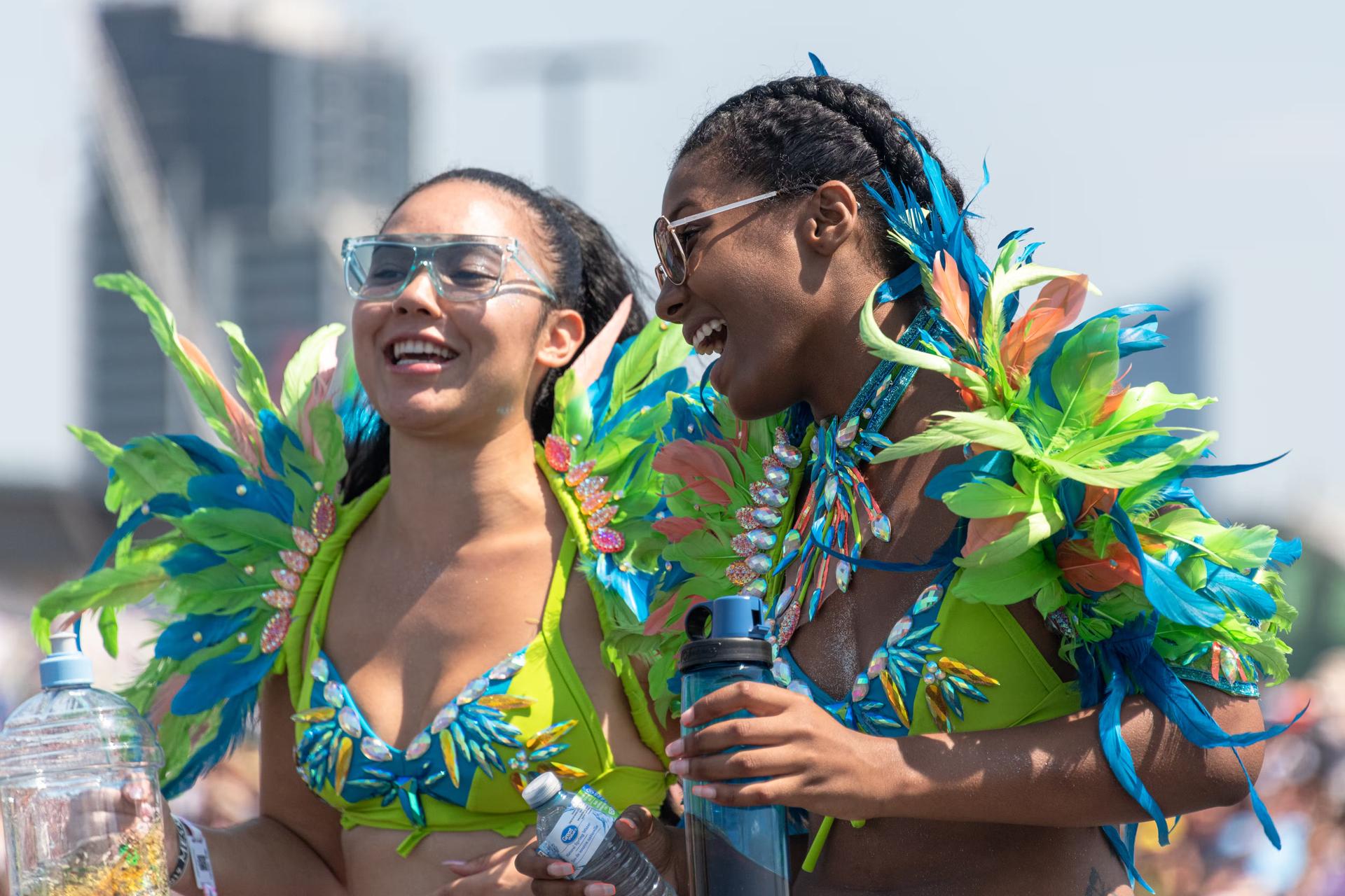 Två unga kvinnor i klargröna och blå fjäderklädda kostymer ler mot varandra vid ett evenemang i Toronto. 