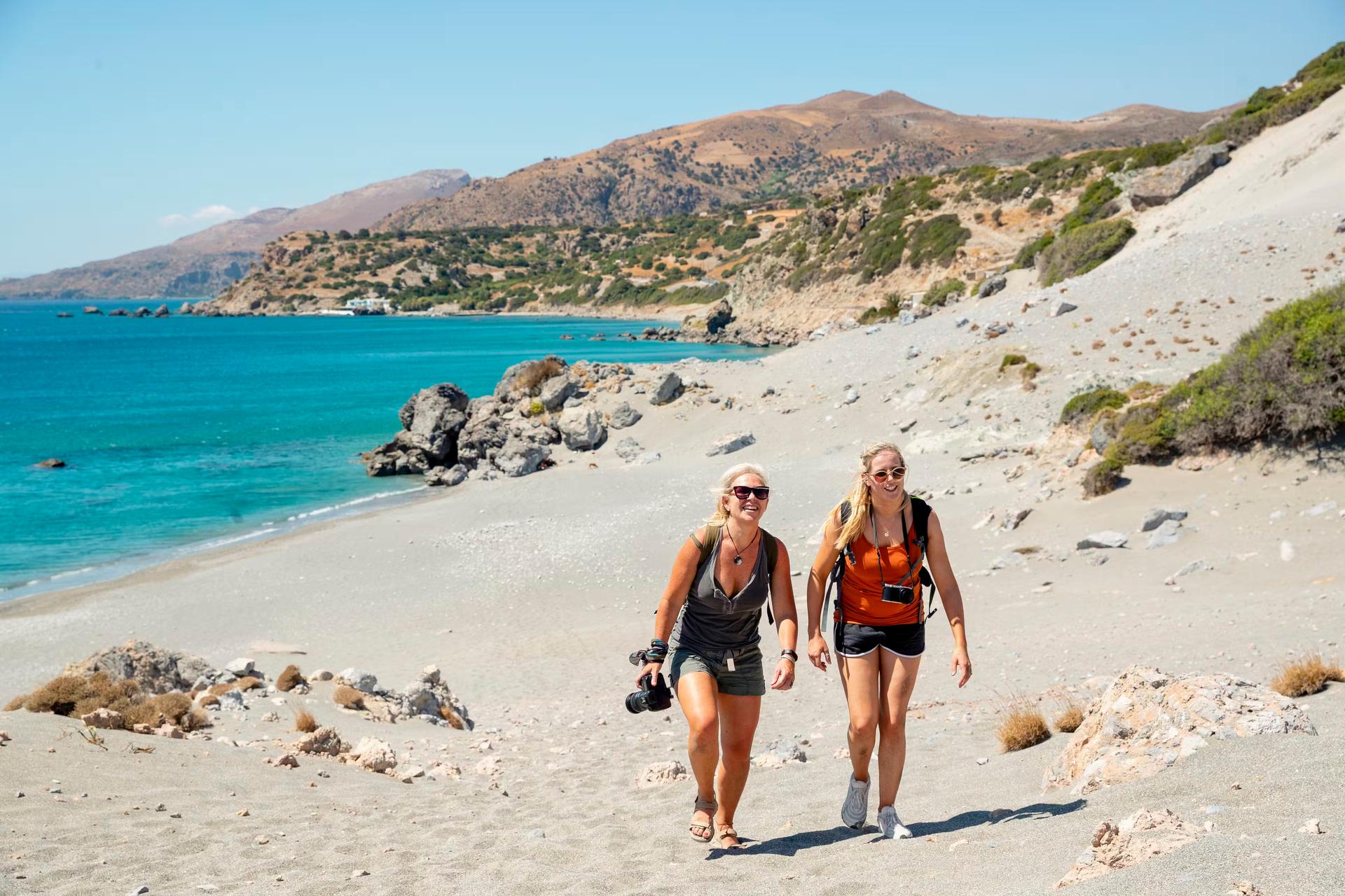 Två blonda kvinnor går på stranden på Kreta.  De ser glada och sorglösa ut i solen och håller kameror.