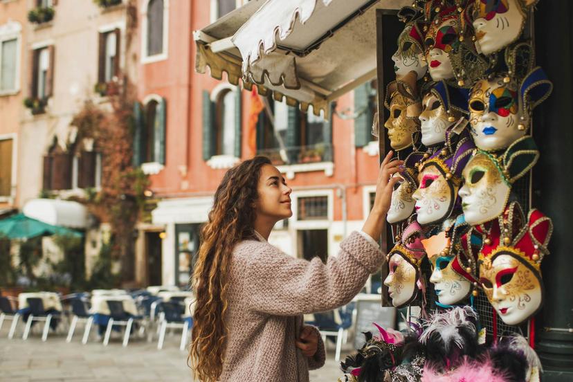 Vacations and shopping in Venice, Italy. Tourist woman want to choose and buy one. Traditional beautiful handmade carnival masks as souvenir selling on street kiosk. Giel enjoying and smiling.