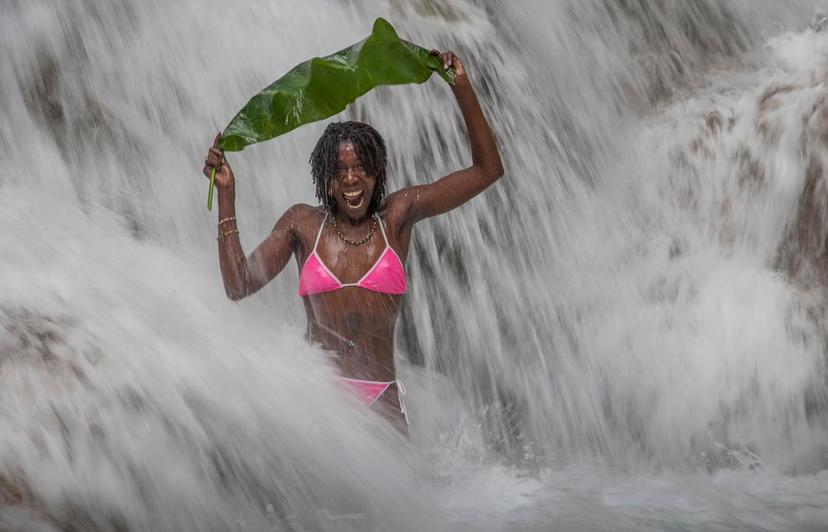 Jamaica. Echo Rios. Young smiling jamaican woman with pink bikini in the middle of the world famous Dunns River Falls, covering herself with a banana leaf.