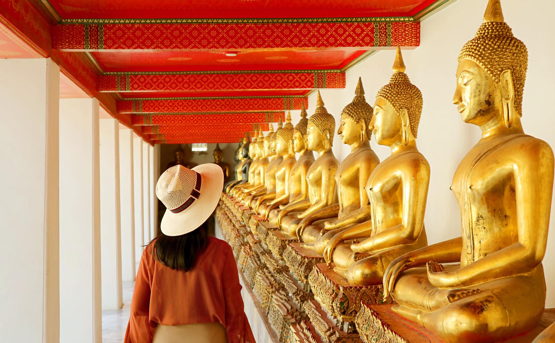En kvinna som bär kläder som täcker hennes axlar går längs en vägg av gyllene buddhor i ett thailändskt tempel