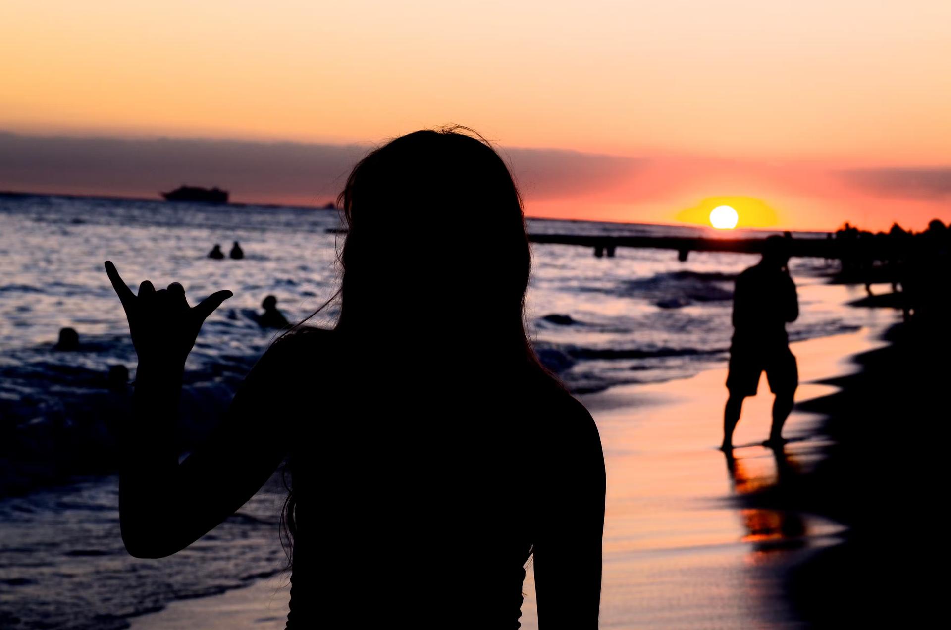 A female in silhouette as the sun sets over the beach holds up her hand in a hang loose sign or 