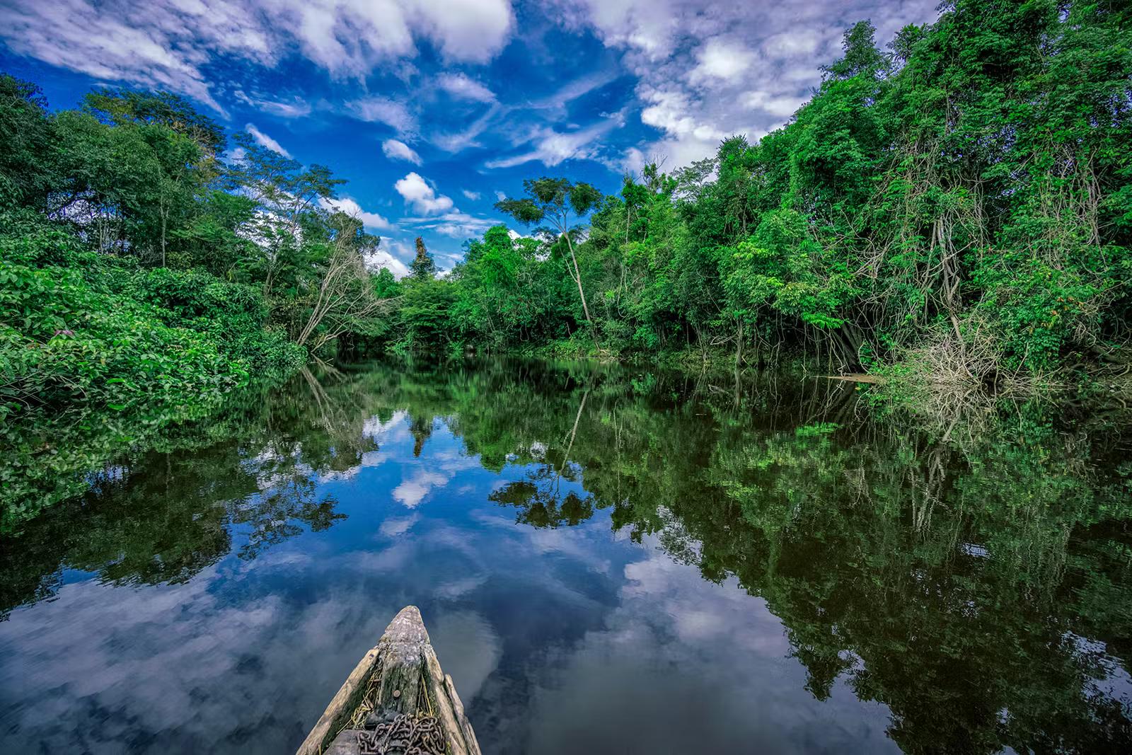 En träkanot driver nerför en glasig Nanay-flod med Amazonasskog på båda stränderna.  Fotot är taget från kanotens kropp, med bara spetsen i ram.  Den molniga blå himlen reflekteras i det stilla vattnet.
