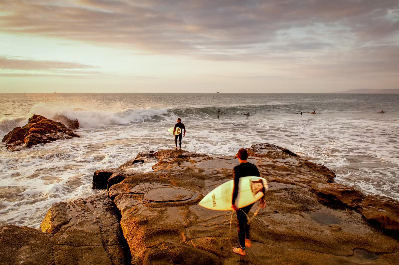 Två surfare går ut på en rostfärgad sten mot bränningen vid solnedgången.  Det finns flera andra surfare redan i havet.
