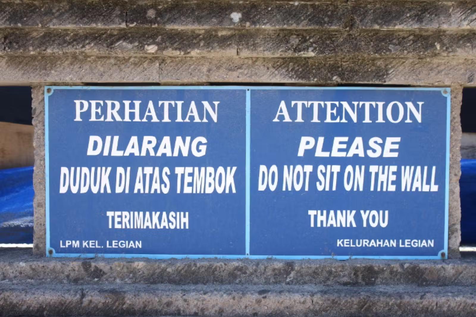 A pair of signs, one in Indonesian and the other in English, warning travellers to not sit on a wall in Bali. 