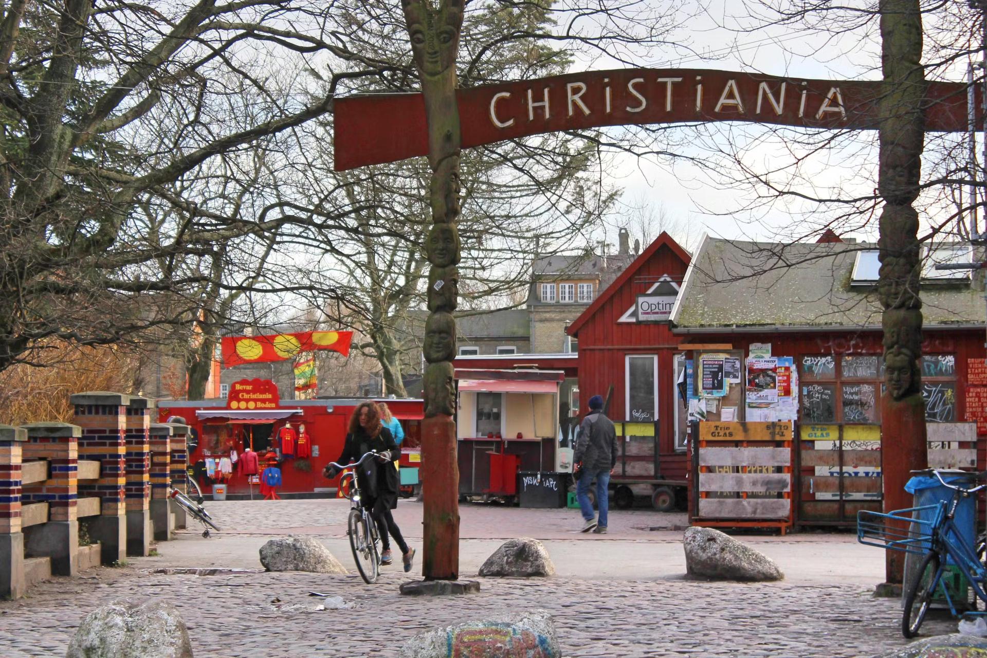 Copenhagen neighbourhoods - A woman cycles by the large wooden Christiania sign. In the background we can see wooden huts and stalls, more bicycles and one other person wearing a hat and warm coat. 