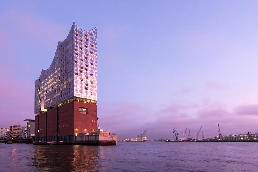 The exterior of Elbphilharmonie (Elbe Philharmonic Hall) in the HafenCity of Hamburg. The photo is taken from the water at dusk. The reflective glass exterior of the concert hall sits on top of a red-brick structure.