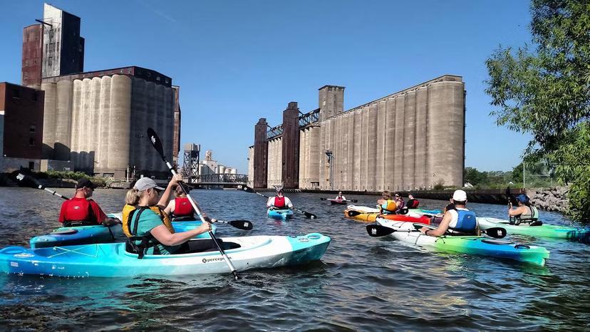 Several kayakers paddle past large concrete grain silos on the Buffalo River