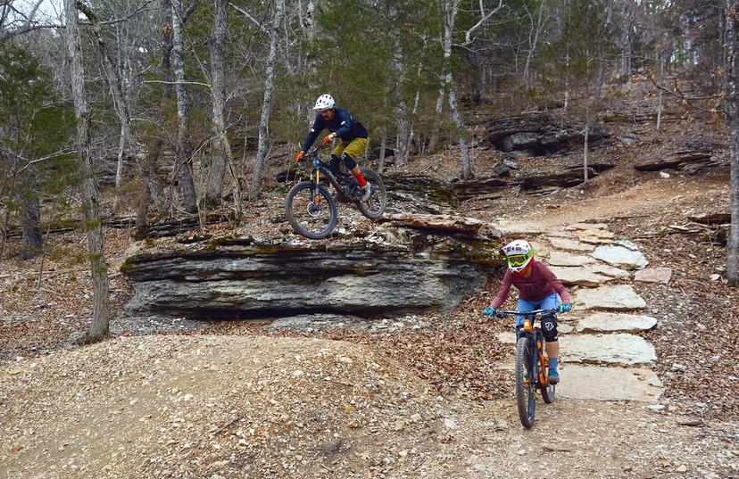 A mountain biker leaps off a rocky outcropping while a second rider takes a side option of a curving rock path