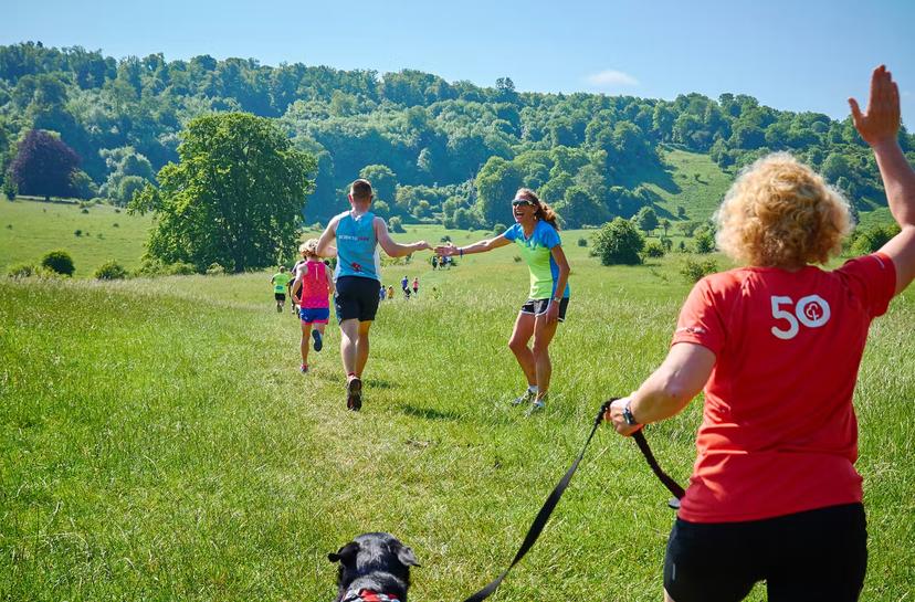 A child and youth run through green grasslands towards a forested hill; a supporter holds her hand out to the youth to slap as he passes, as a runner in the foreground heads away from the camera with her dog on a lead