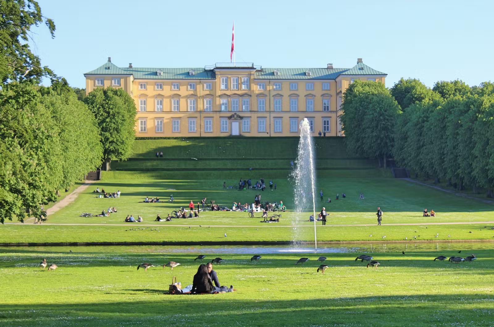 Grupper av människor sitter utspridda längs den vidsträckta gräsmattan och fontänen framför Frederiksbergs slott vid Frederiksberg Have.  Trädgården ligger på 32 hektar mark. 