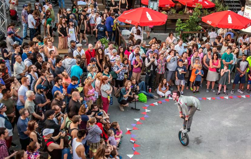 An acrobat stands on a unicycle in a funny pose as a crowd of spectators surround him, sitting on the ground