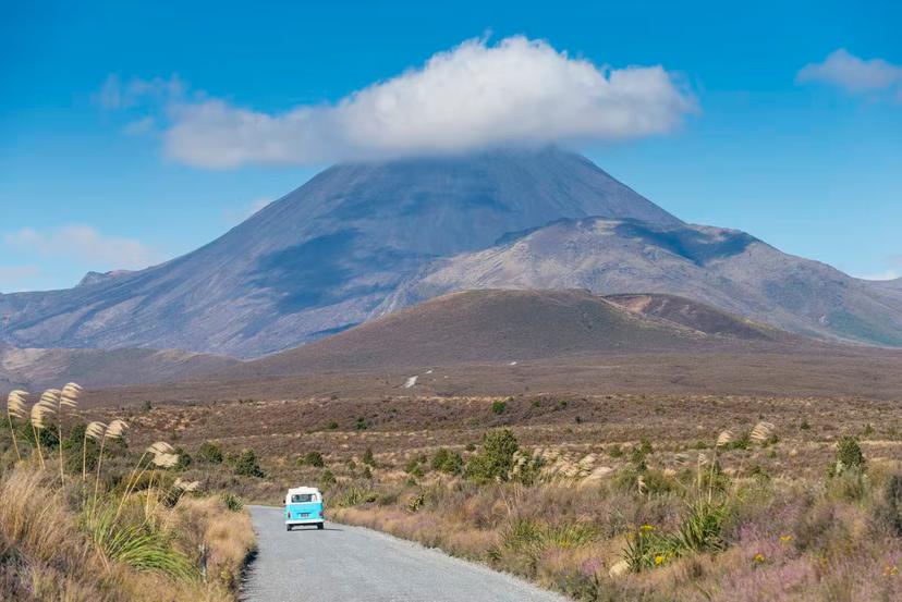 Campervan on road in Tongariro National Park, with volcanic peak (with its rocky summit cloaked in a petite cloud, in background