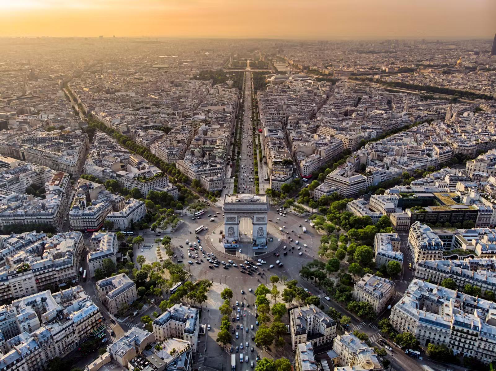 An aerial view over the ornate Arc de Triomphe at sunrise; it sits in the middle of a tree-lined roundabout, with roads radiating out in each direction.