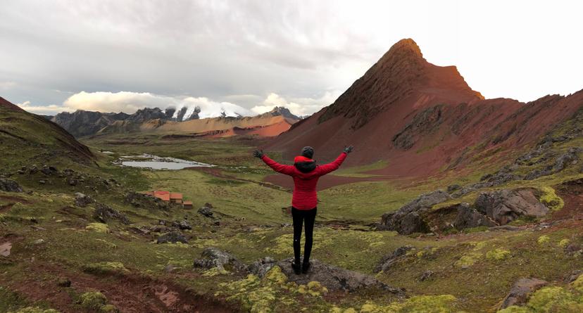A woman in a red and black jacket extends her arms as she looks over a vast mountain landscape. 