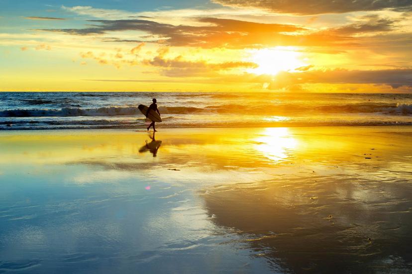 Silhouette of a surfer walking on a sandy beach with a surfboard during sunset. 