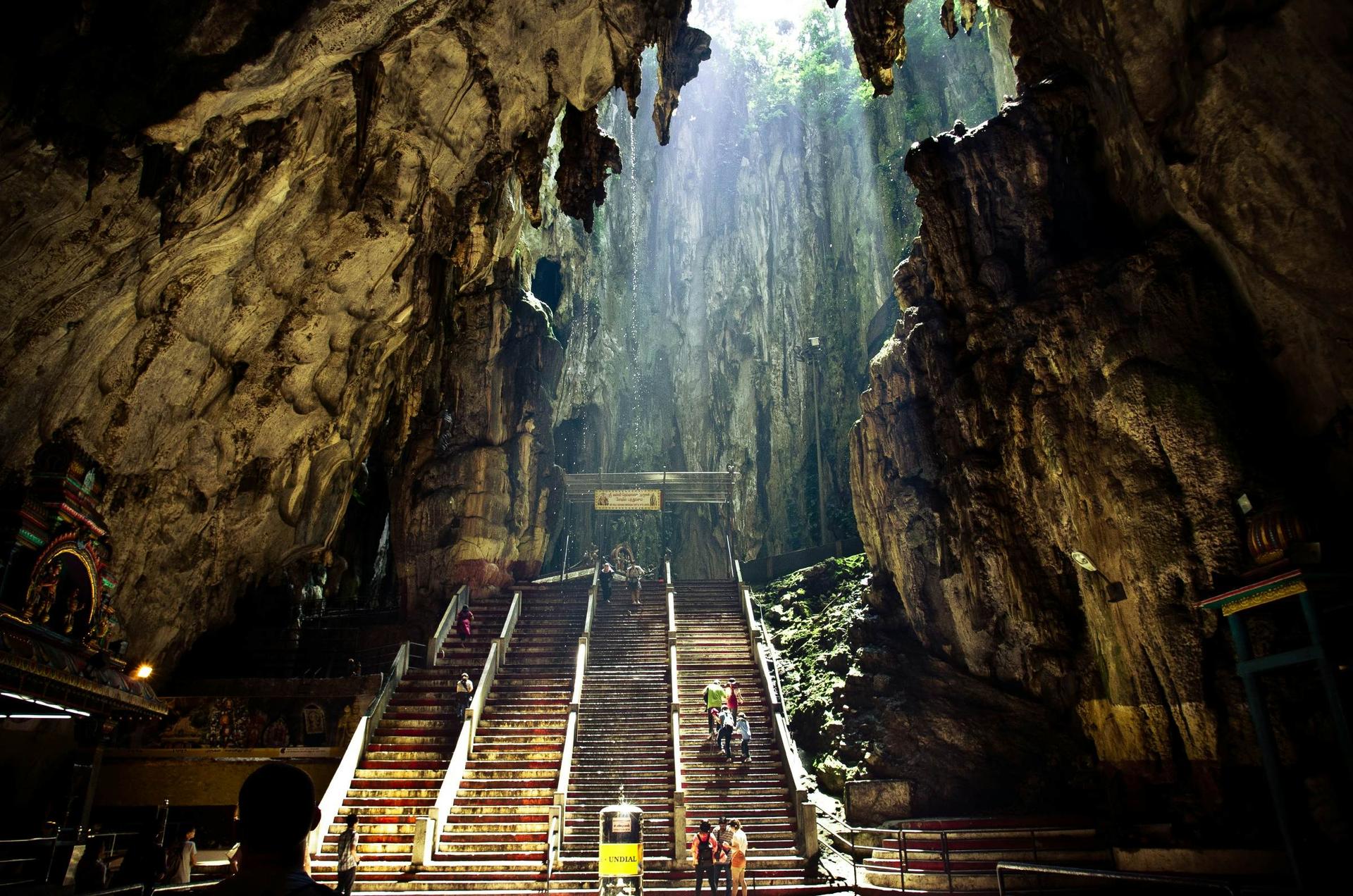 The interior of the Batu Caves near Kuala Lumpur in Malaysia. There are stairs leading further into the cave, while the walls consist of jagged rock. The roof is open, and light streams down from above.
