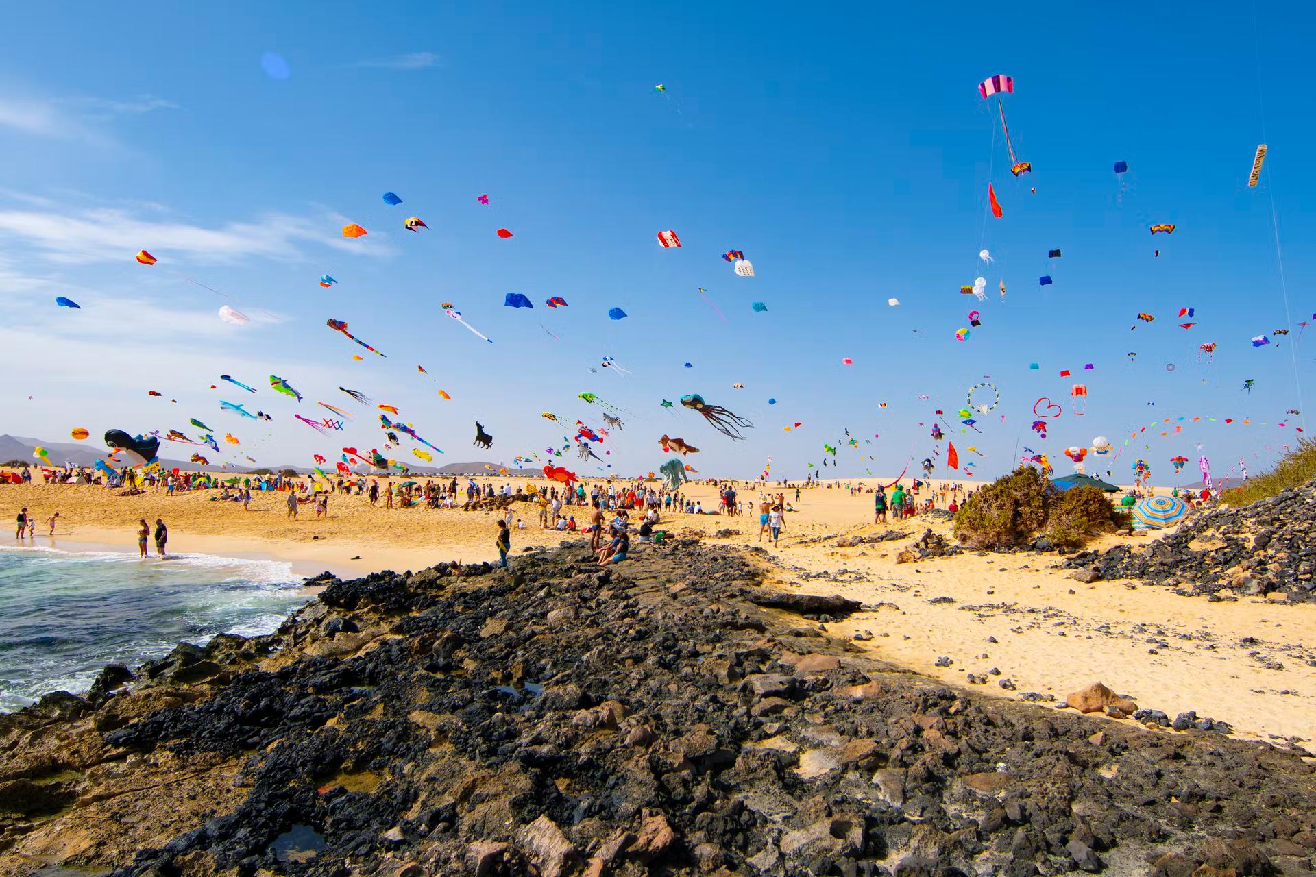 Hundreds of colourful kites being flown on the sandy beach on a windy day; there is a rocky outcrop in the foreground.