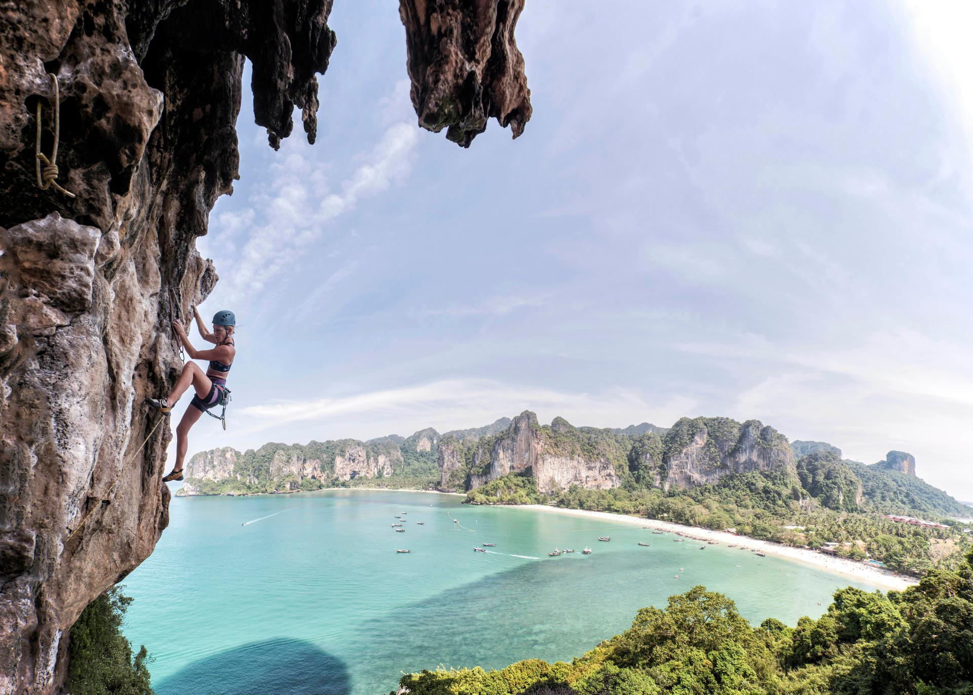 A rock climber scales a sheer rock face in Krabi, Thailand. In the background is a sweeping view of a white-sand beach and turquoise sea.