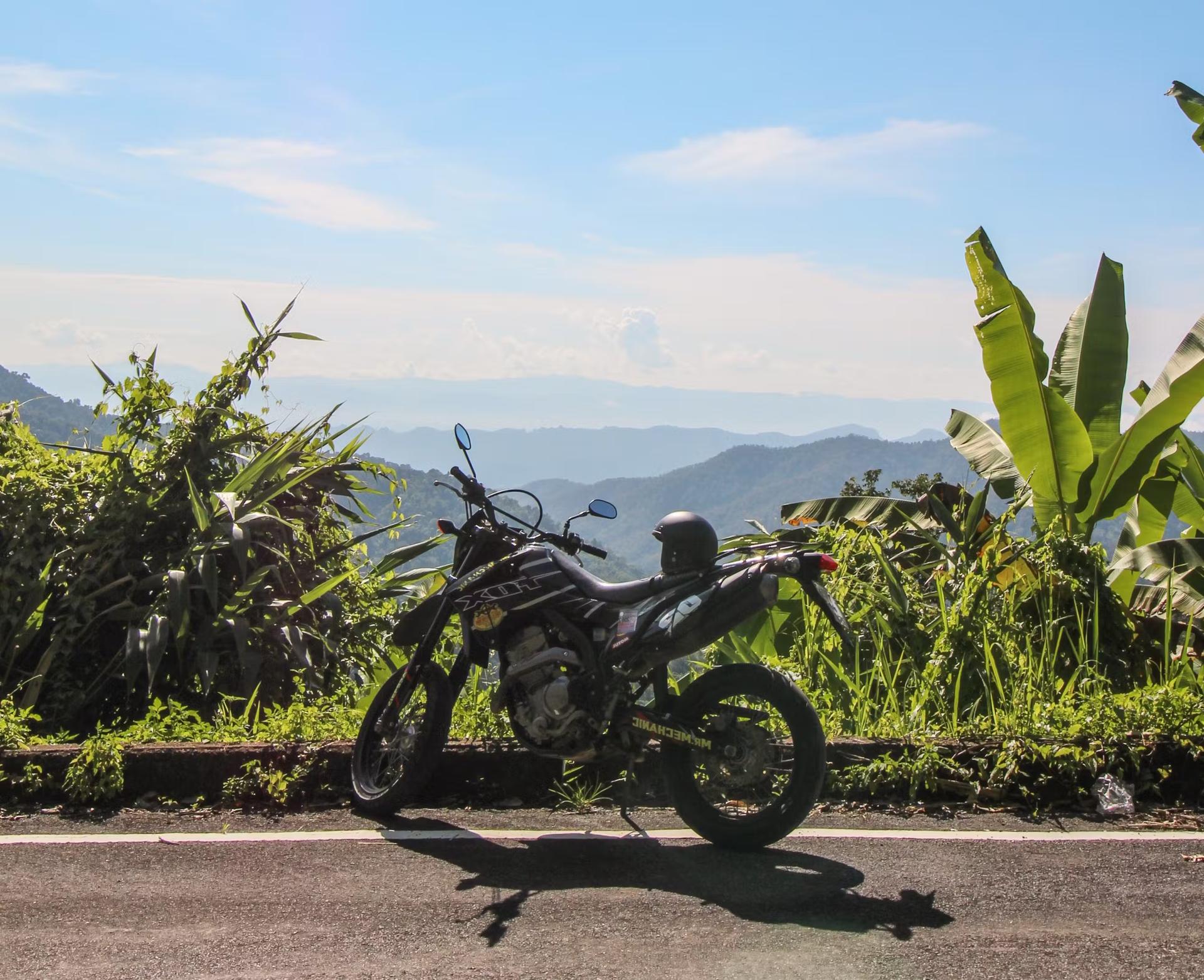 En motorcykel står parkerad vid en naturskön utsiktspunkt längs Samoeng-slingan nära Chiang Mai.  En hjälm sitter på cykelsätet medan utsikten över gröna dalar är synliga bortom.