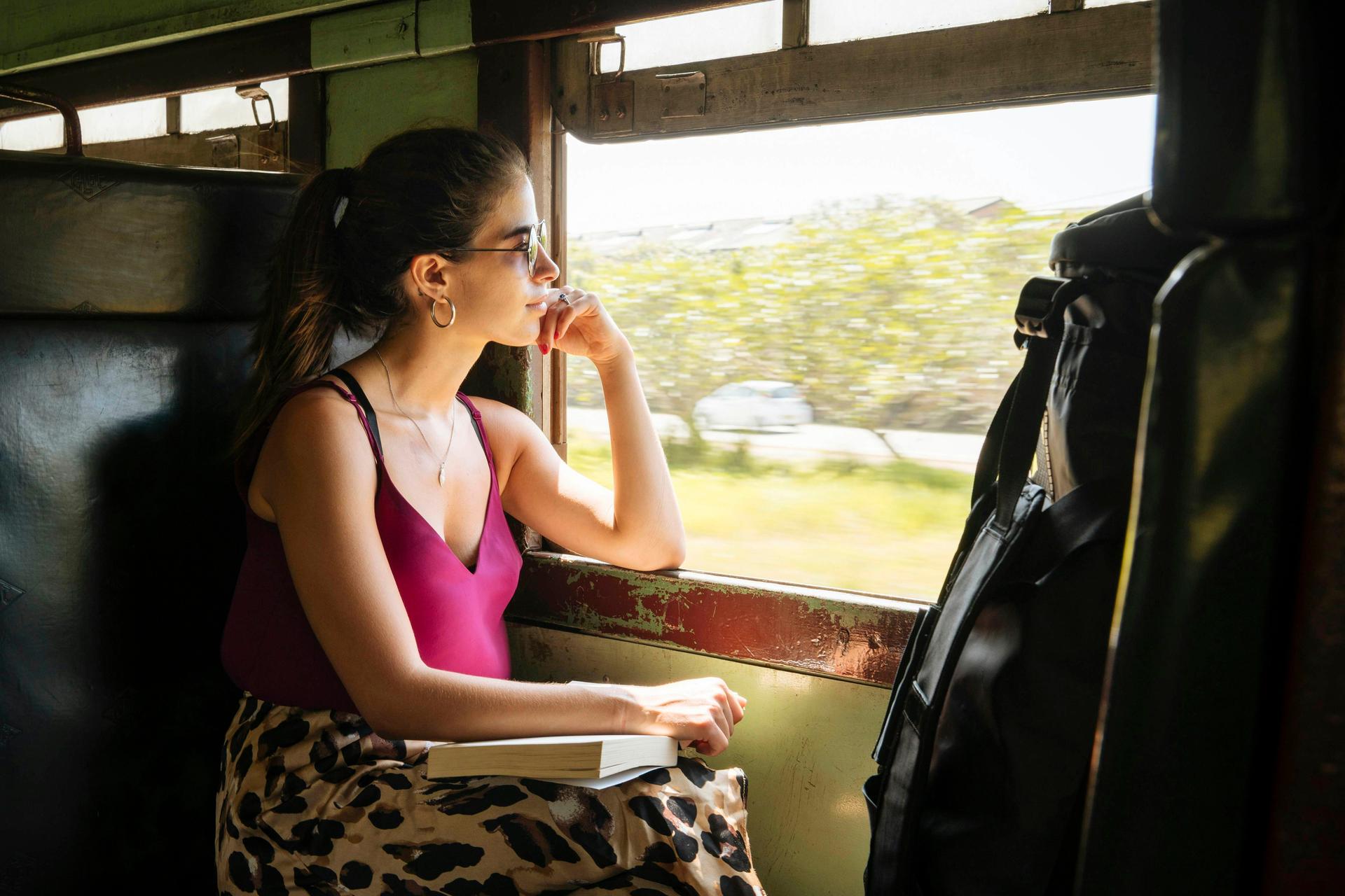 A girl wearing sunglasses sits in a window seat on a Sri Lankan train from Colombo to Galle. A book rests on her lap as she stares out of the open window, where green countryside is visible.