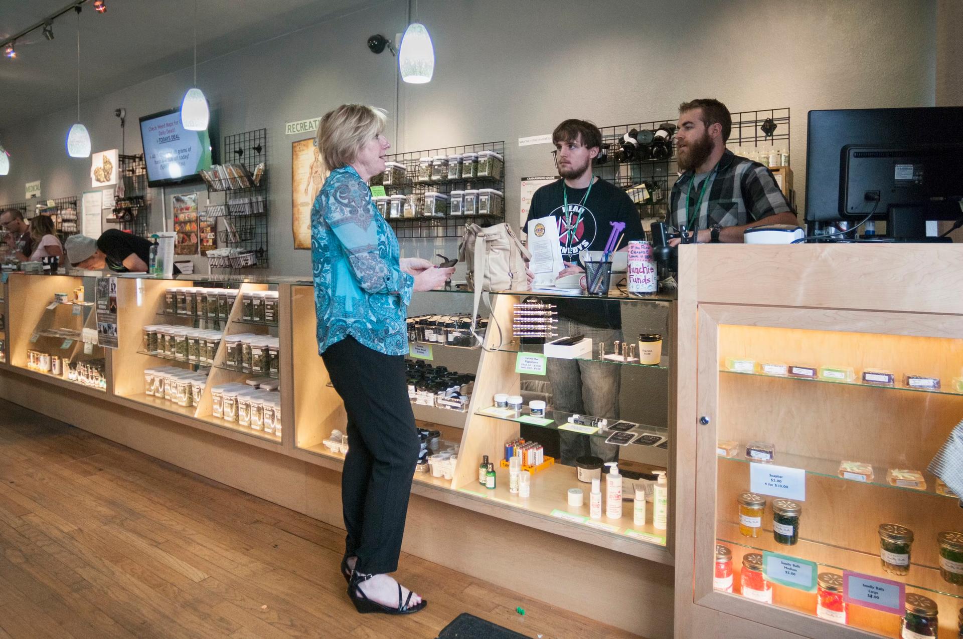 A middle aged white woman with short blond hair in a blue floral tunic, black slacks, and black sandals speaks with two budtenders at a Denver, Colorado cannabis dispensary. A long display case and counter made of blonde wood and glass shows cases a variety of edible and topical cannabis products and concentrates, while jars of flower sit on shelves on the wall
