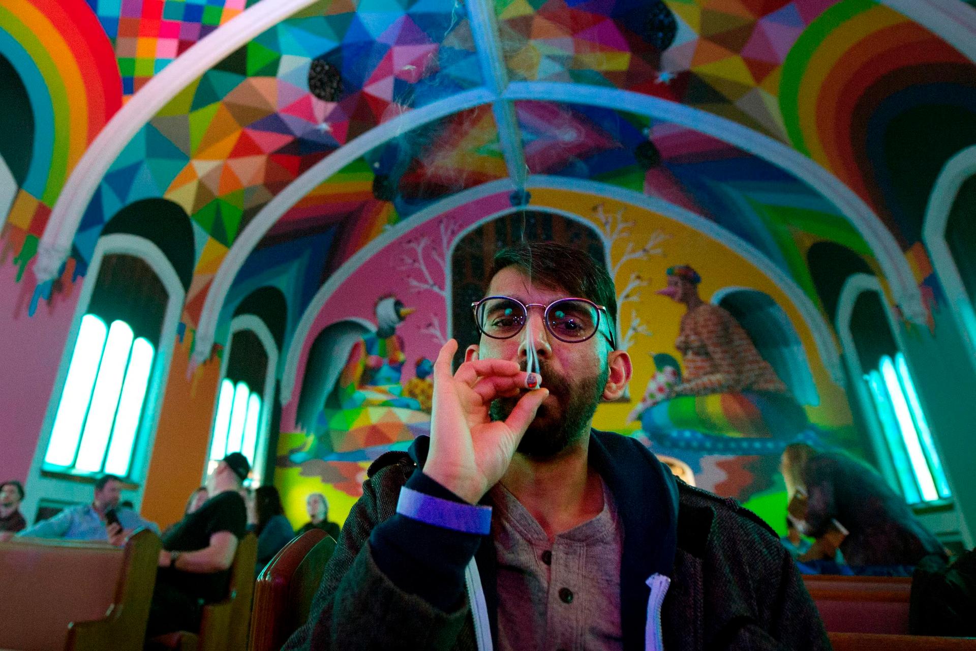 A man with glasses smokes a joint while gazing slightly down at the viewer. He is framed by the rainbow arched ceiling of the International Churhc of Cannabis. Behind him are other people sitting in pews for an Elevationist non denominational service
